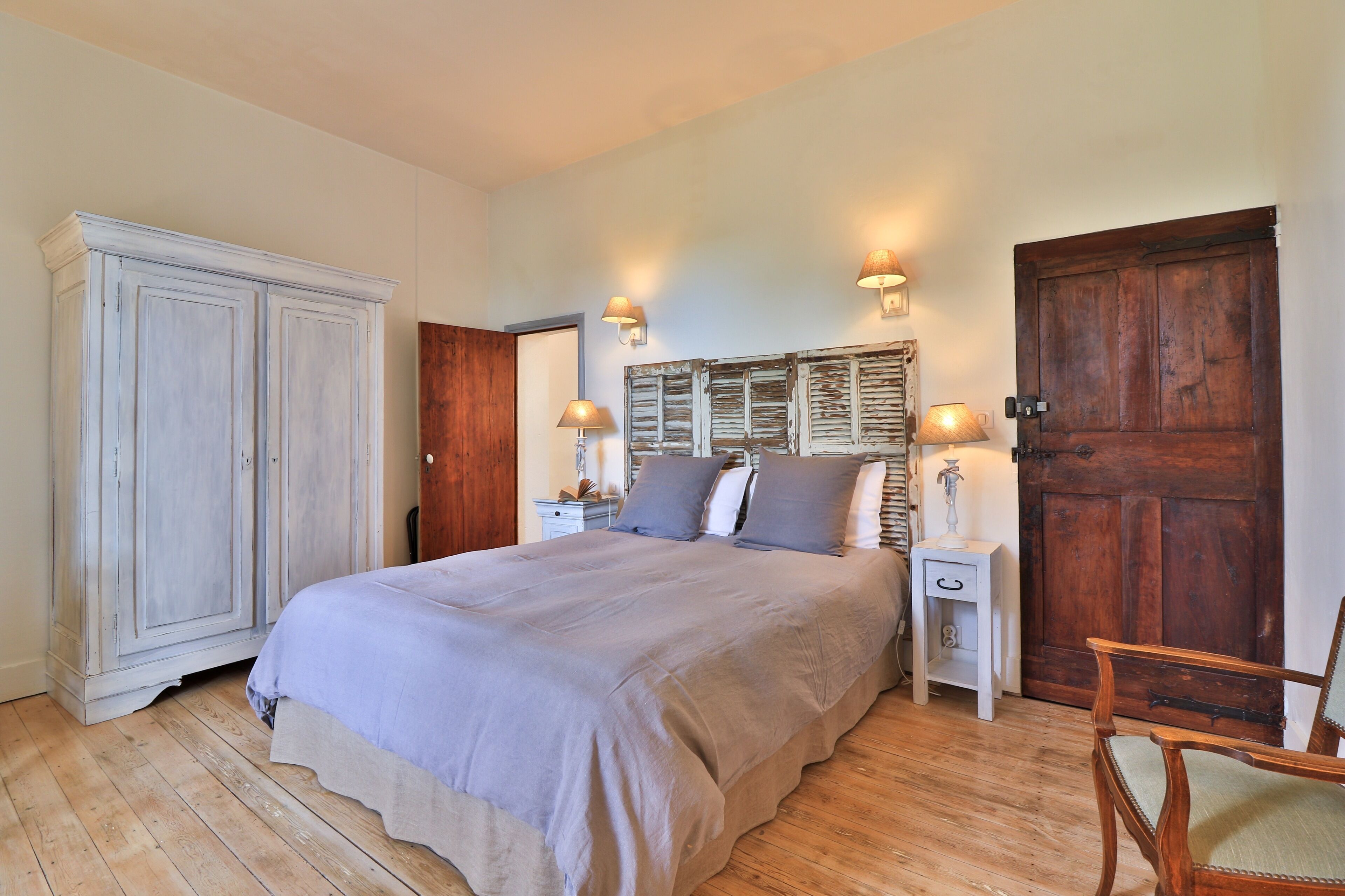 A bedroom with a large bed, wooden wardrobe, two bedside tables with lamps, and a wooden door.