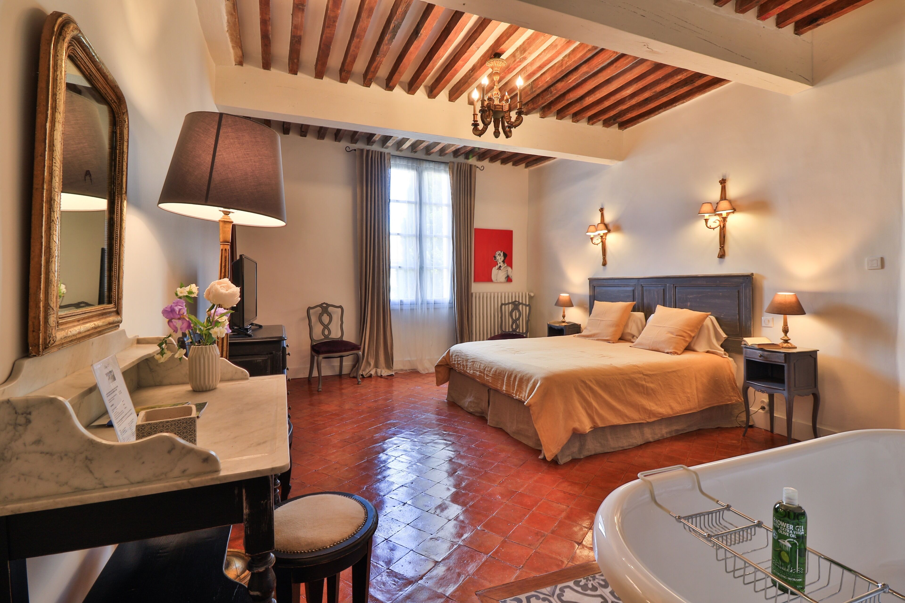 A hotel room with a bed, a desk, a chair, a desk lamp, a mirror, a chandelier, a window with curtains, and a bathtub.
