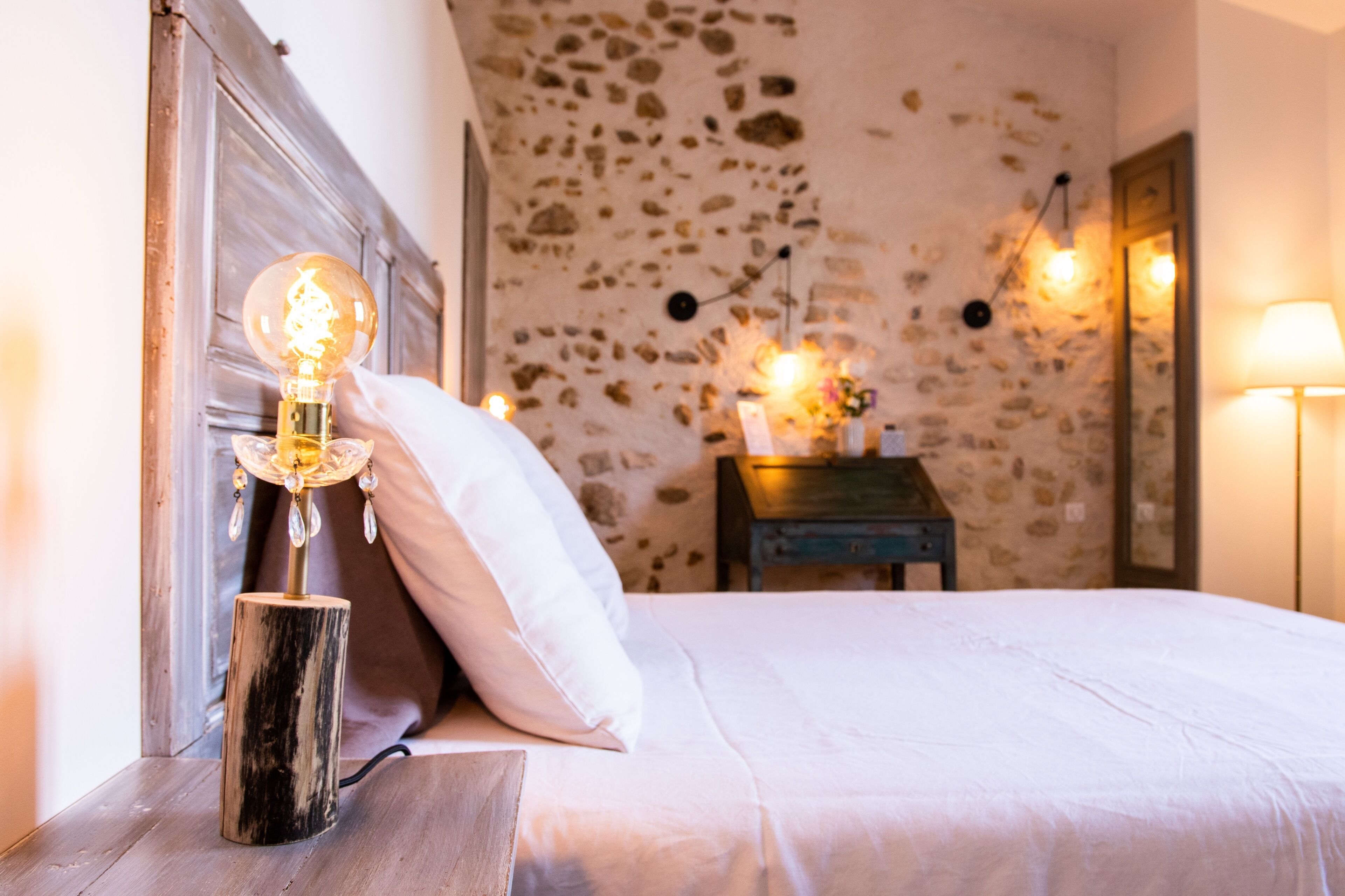 A bedroom with a stone wall, a wooden bedside table with a lamp, a white bed, and a nightstand with a lamp.