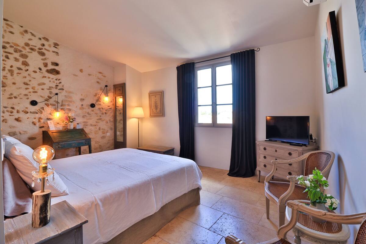 Een slaapkamer met een stenen muur, een bed, een bedlamp, een televisie, een stoel en een dressoir.