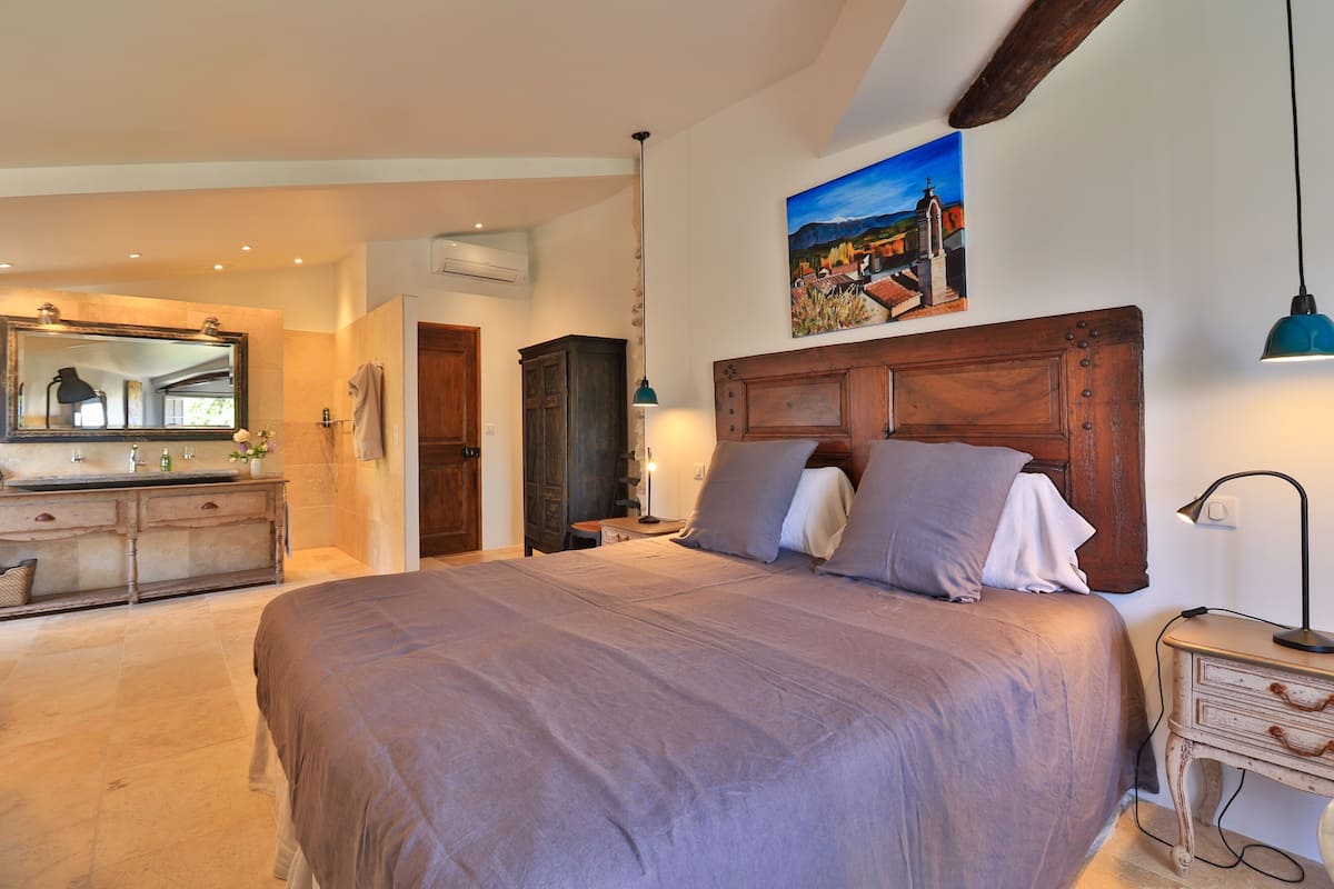 Een slaapkamer met een groot bed, een houten hoofdbord, een schilderij aan de muur, en een badkamer met spiegel en wastafel.