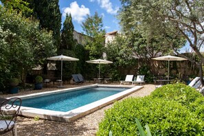 Una piscina rectangular con borde de azulejos, rodeada de grava. Hay sillones de descanso y sombrillas, además de una mesa con sillas.
