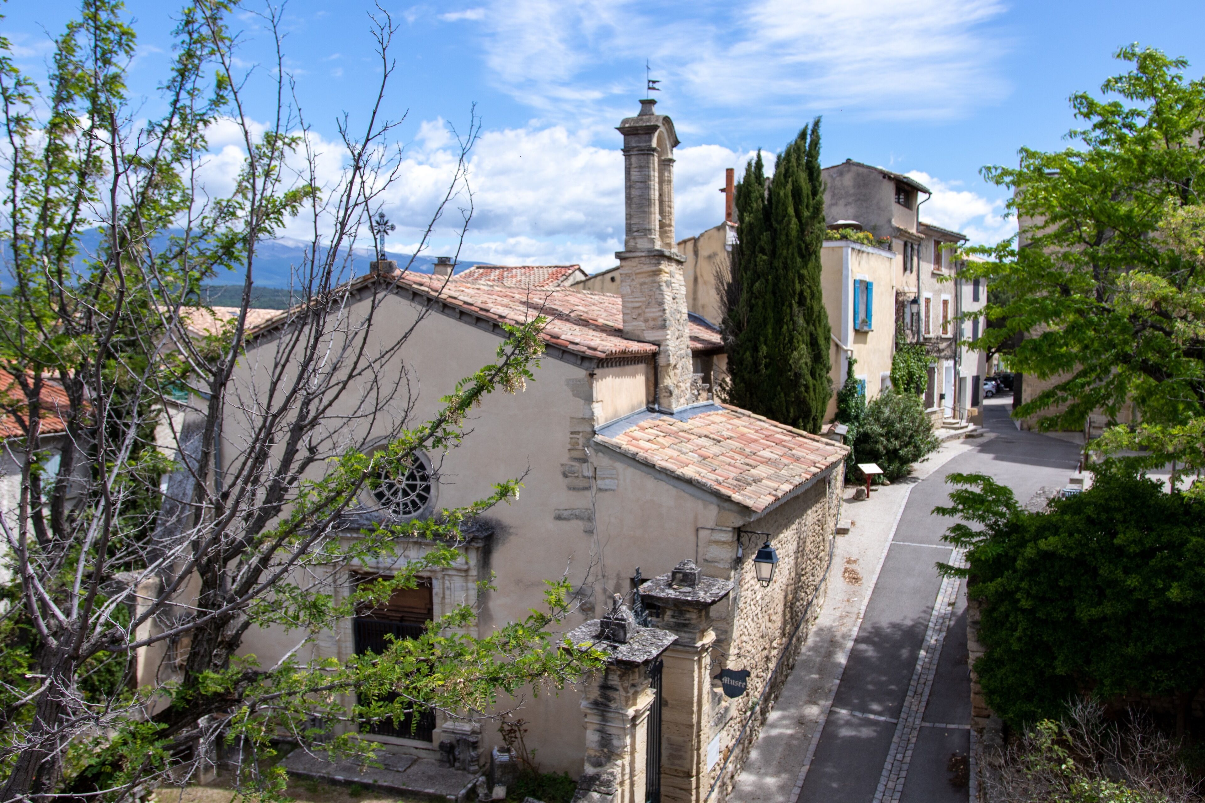 Une rue étroite bordée de bâtiments traditionnels en pierre, une église avec un clocher et, au loin, une vue sur la mer.