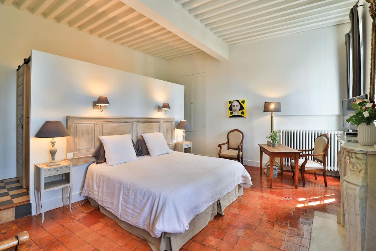 Een slaapkamer met een groot bed, twee nachtkastjes met lampen, een stoel, een klein tafeltje met een plant en een radiator.