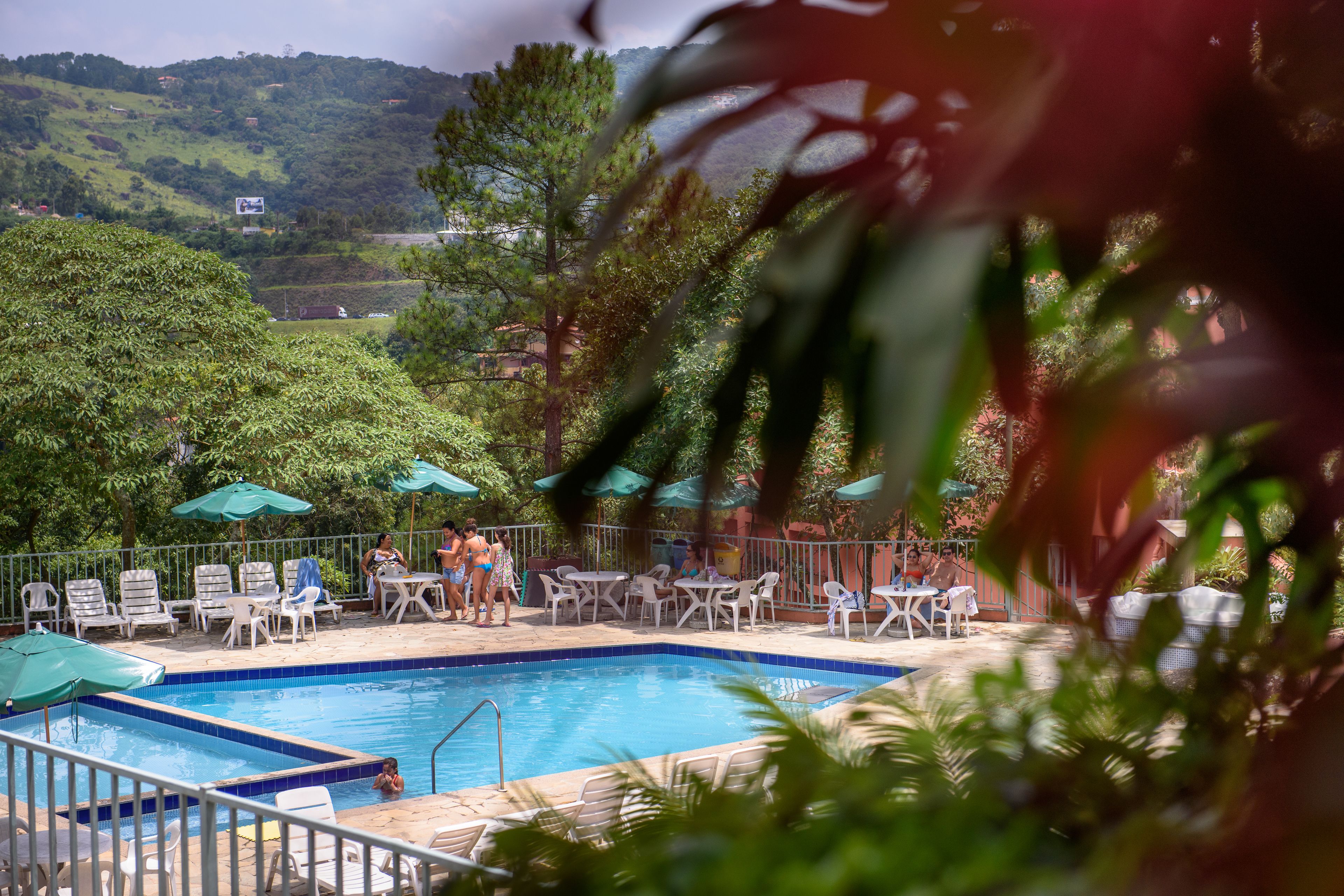 A pool area with lounge chairs and umbrellas, surrounded by greenery and hills in the background.