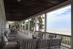 A covered deck with outdoor seating, a view of the beach, and palm trees.