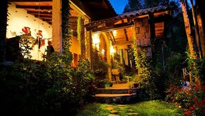 A rustic wooden house with a stone facade, a porch with potted plants, and a well-lit garden at night.