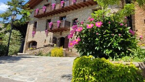 A stone building with a wooden balcony, surrounded by lush greenery and flowering plants. - Mas D'en Roqueta (Montferrer I Castellbo)