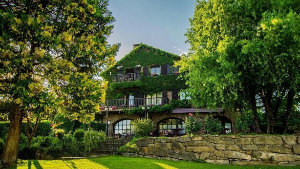 A two-story house with a stone foundation, a green roof, and a balcony with plants. - Mas D'en Roqueta (Montferrer I Castellbo)