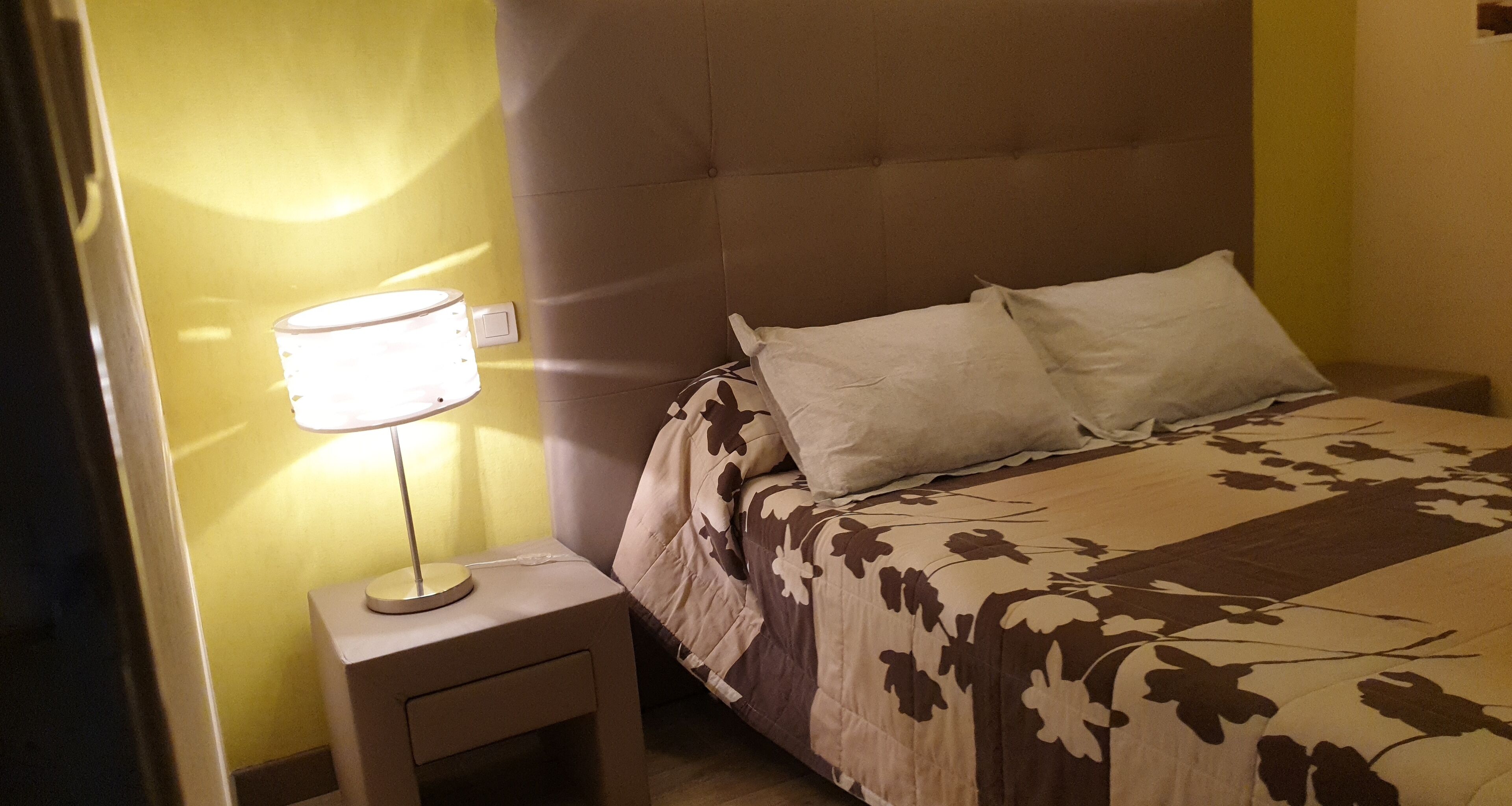 A neatly made bed with a floral-patterned bedspread and two pillows, a bedside lamp on a small table, and a wall switch on the left.
