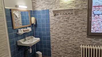 A bathroom with a stone-patterned wall, a blue-tiled wall, a white sink, a mirror, a towel dispenser, and a window with a view of a red roof.