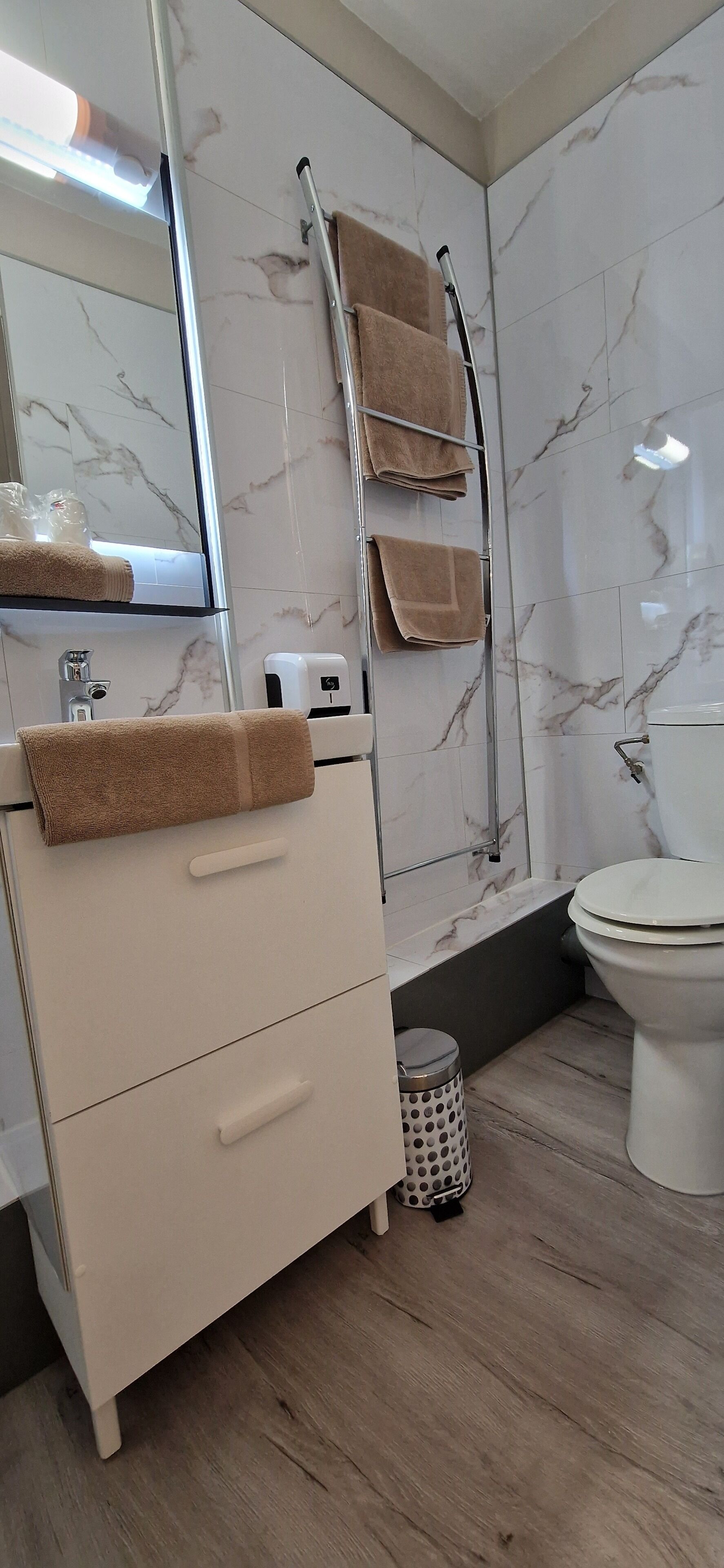 A modern bathroom with a marble backsplash, a white sink with a beige towel, a glass shower enclosure, and a toilet.