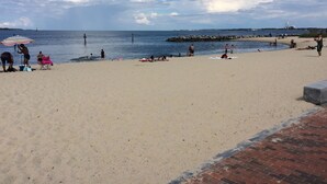 A beach with people sunbathing and swimming, a pier in the background, and a clear sky.