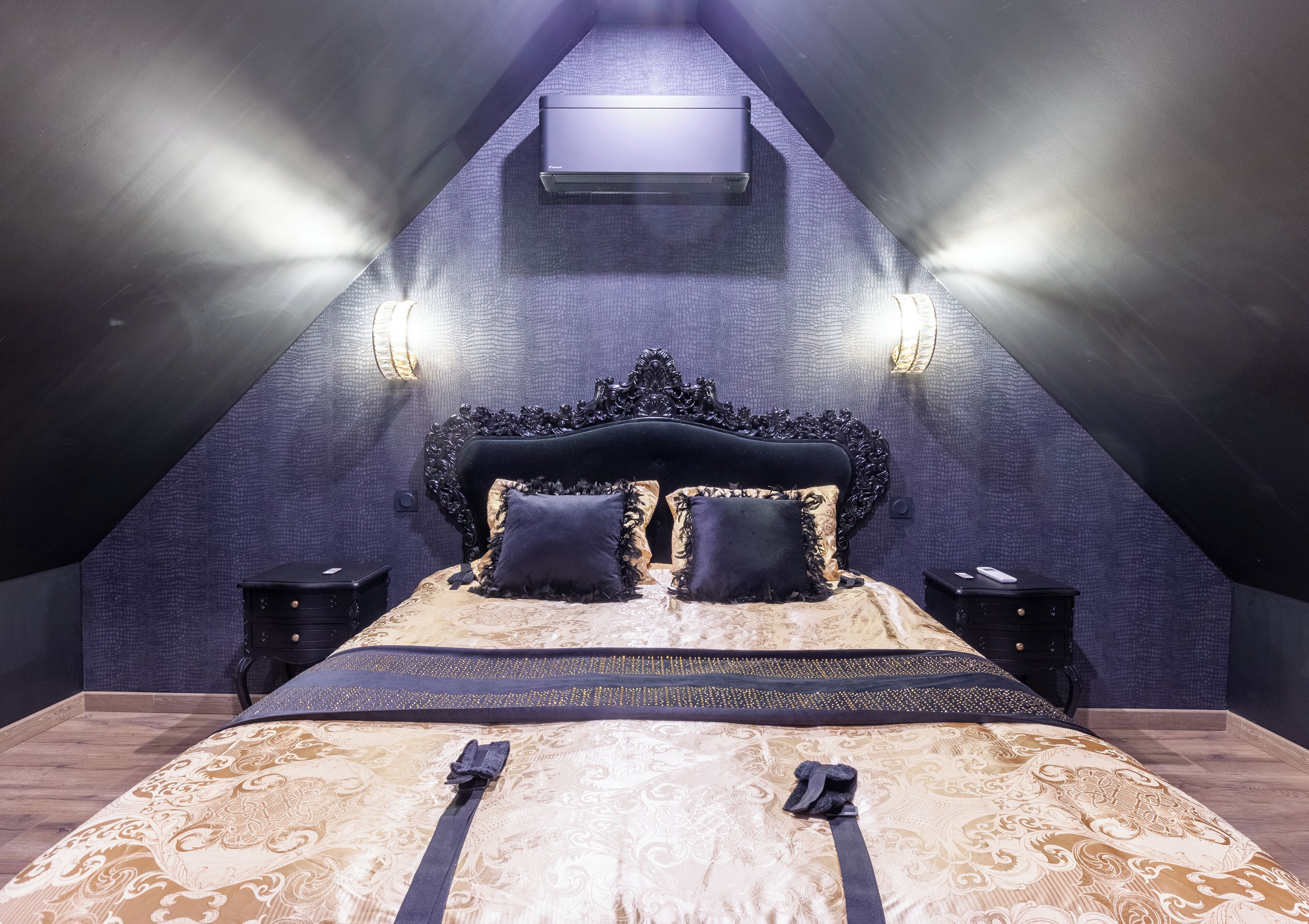 A bedroom with a vaulted ceiling, a large bed with ornate headboard, two bedside tables, and wall-mounted lighting.