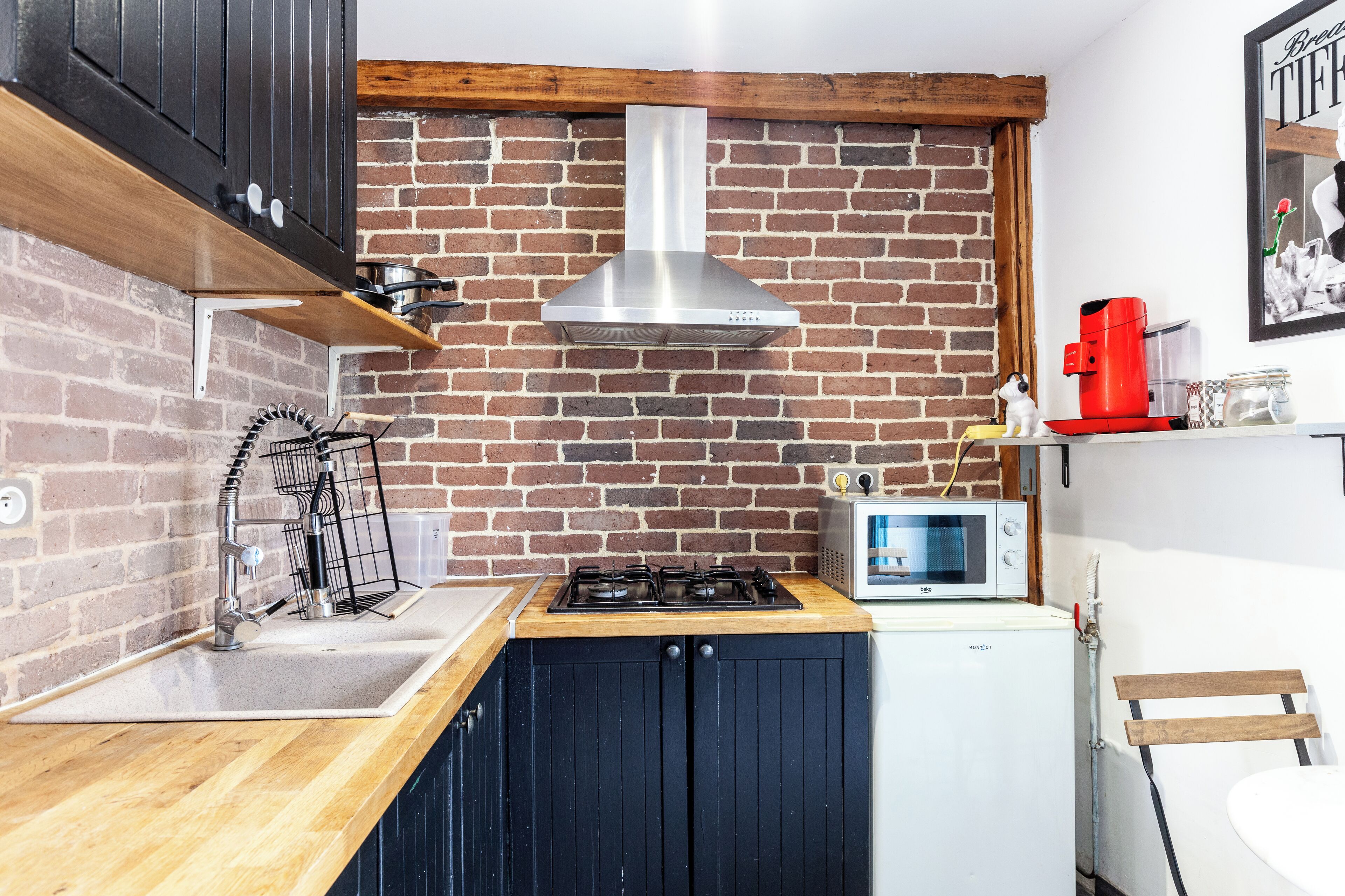 Una cocina con pared de ladrillo, extractor de acero inoxidable, microondas y cafetera roja.