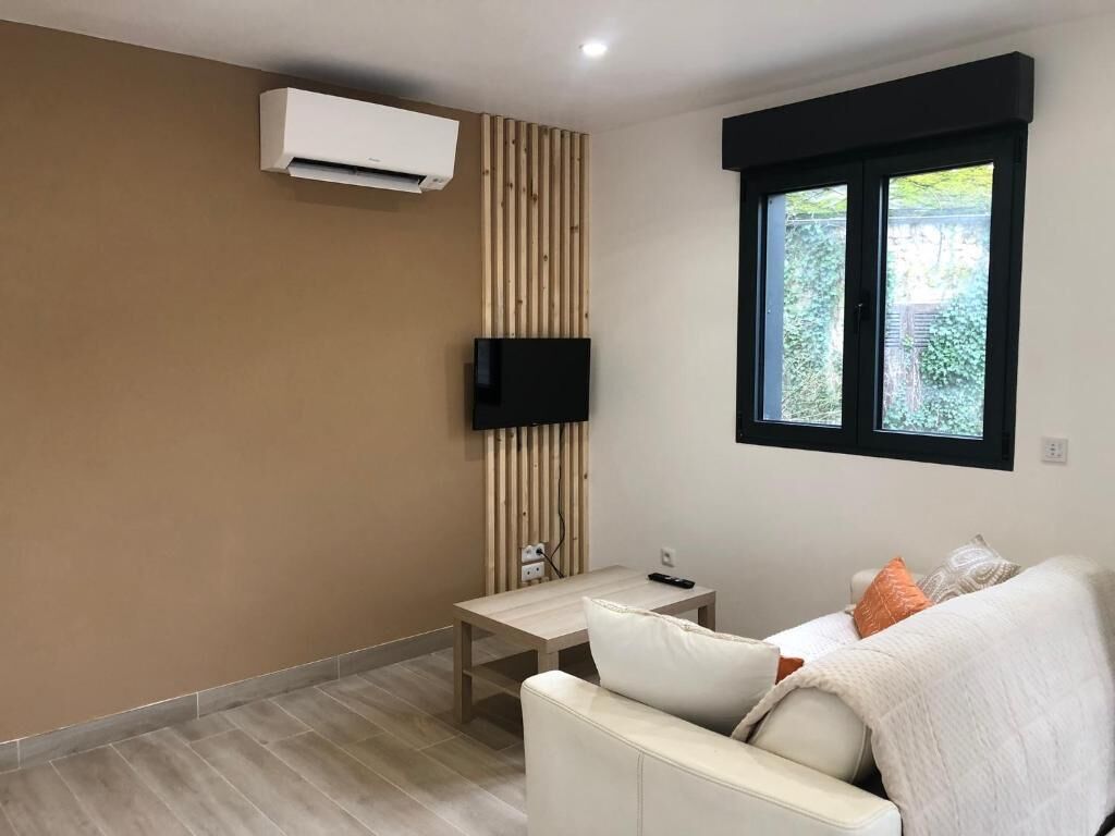 Un salon moderne avec un canapé, un téléviseur fixé au mur, une fenêtre donnant sur un paysage verdoyant et un panneau en bois sur le mur.