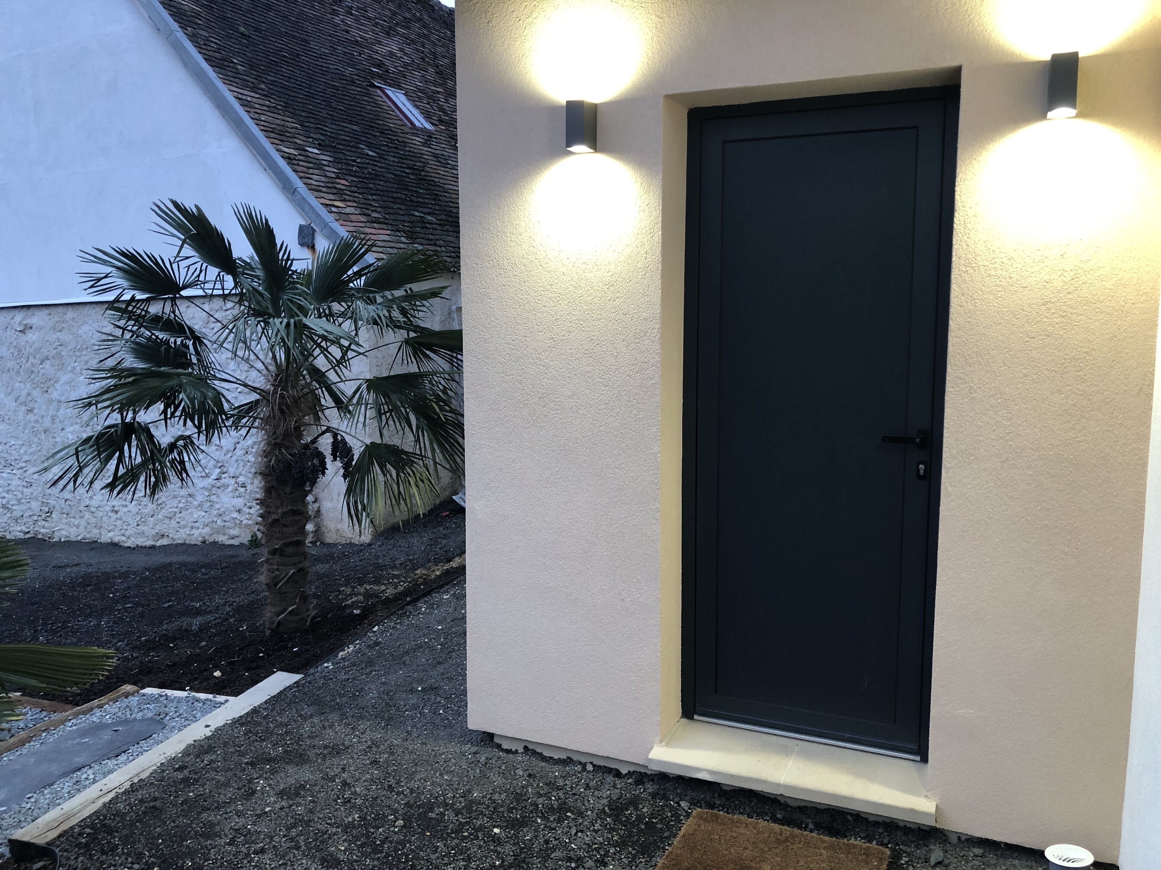 Un bâtiment avec une porte noire, deux lampes fixées au mur et un palmier.