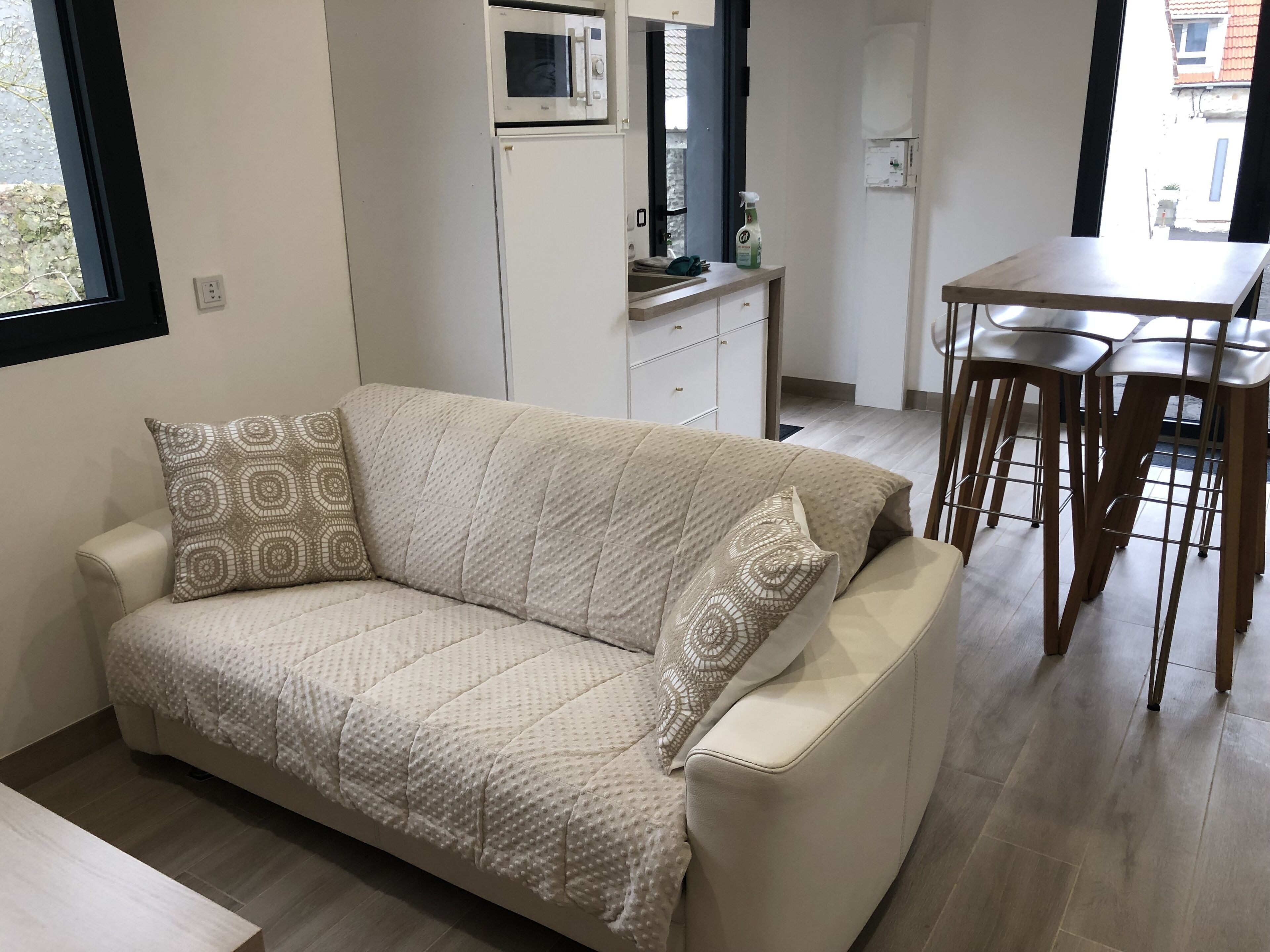 Una sala de estar moderna con un sofá color beige, cojines estampados, una mesa de comedor de madera con taburetes altos, un microondas y una pequeña cocina.