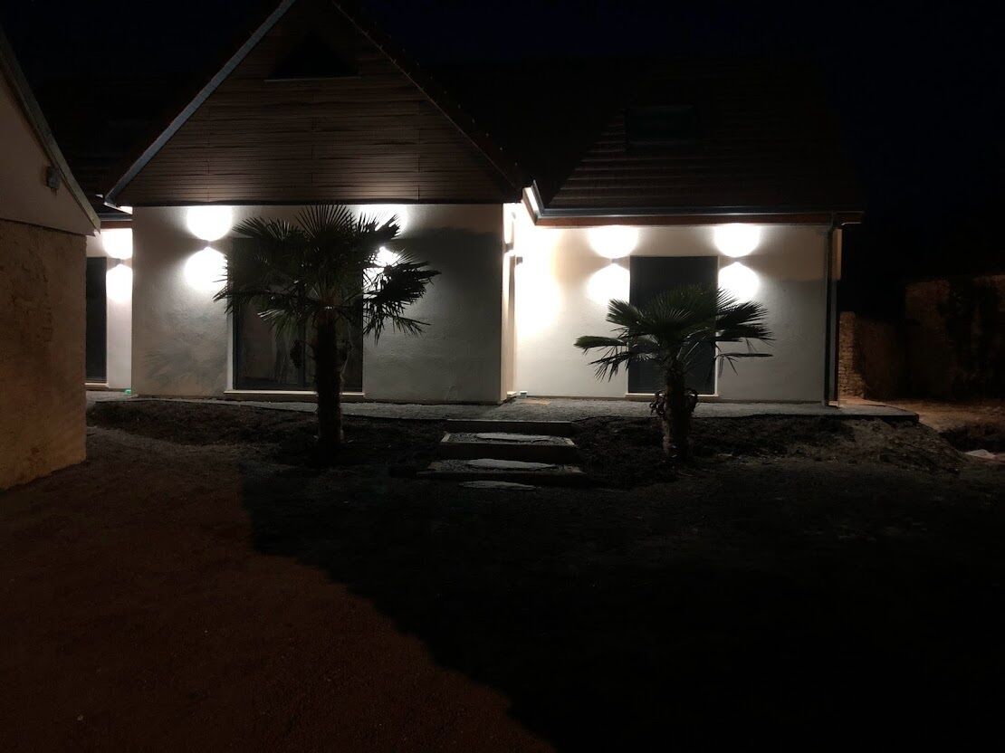 A modern house with a covered porch, two palm trees, and a well-lit exterior at night.