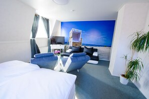 A hotel room with a bed, a blue armchair, a small table with a vase of flowers, a television, and a beach mural on the wall. - Hotel Forsthaus Sellin (Sellin)