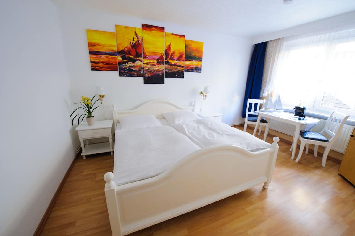 Une chambre d’hôtel avec un lit blanc, un bureau avec une chaise, une petite table avec une lampe et trois tableaux accrochés au mur.