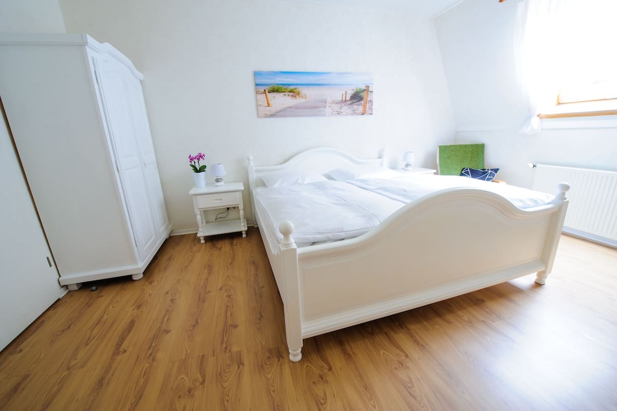 Une chambre à coucher avec un lit blanc, une table de chevet, une commode et un tableau représentant une scène de plage.