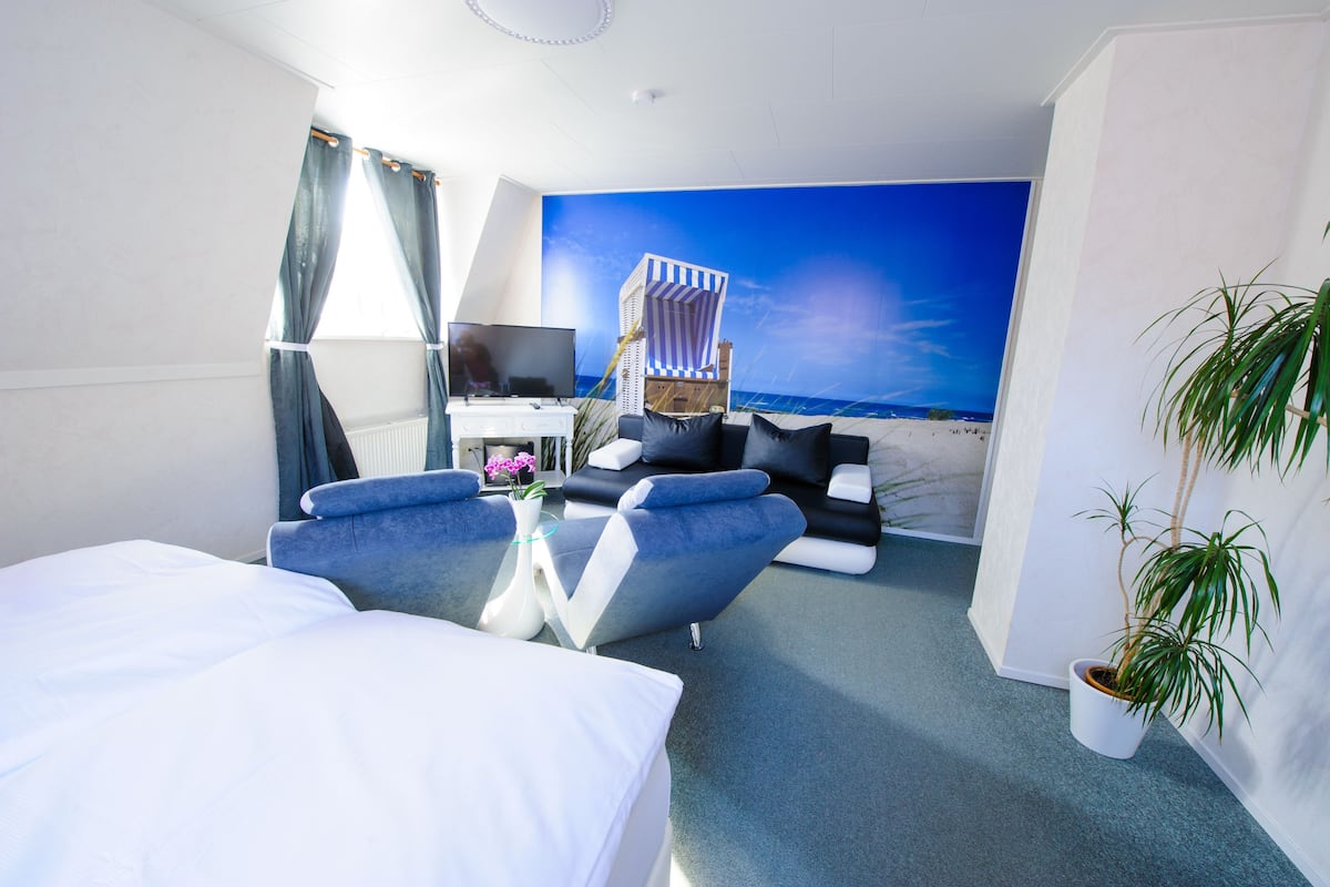 Une chambre d’hôtel avec un lit, un coin salon doté d’une télévision, une fresque murale sur le thème de la plage et une plante en pot.