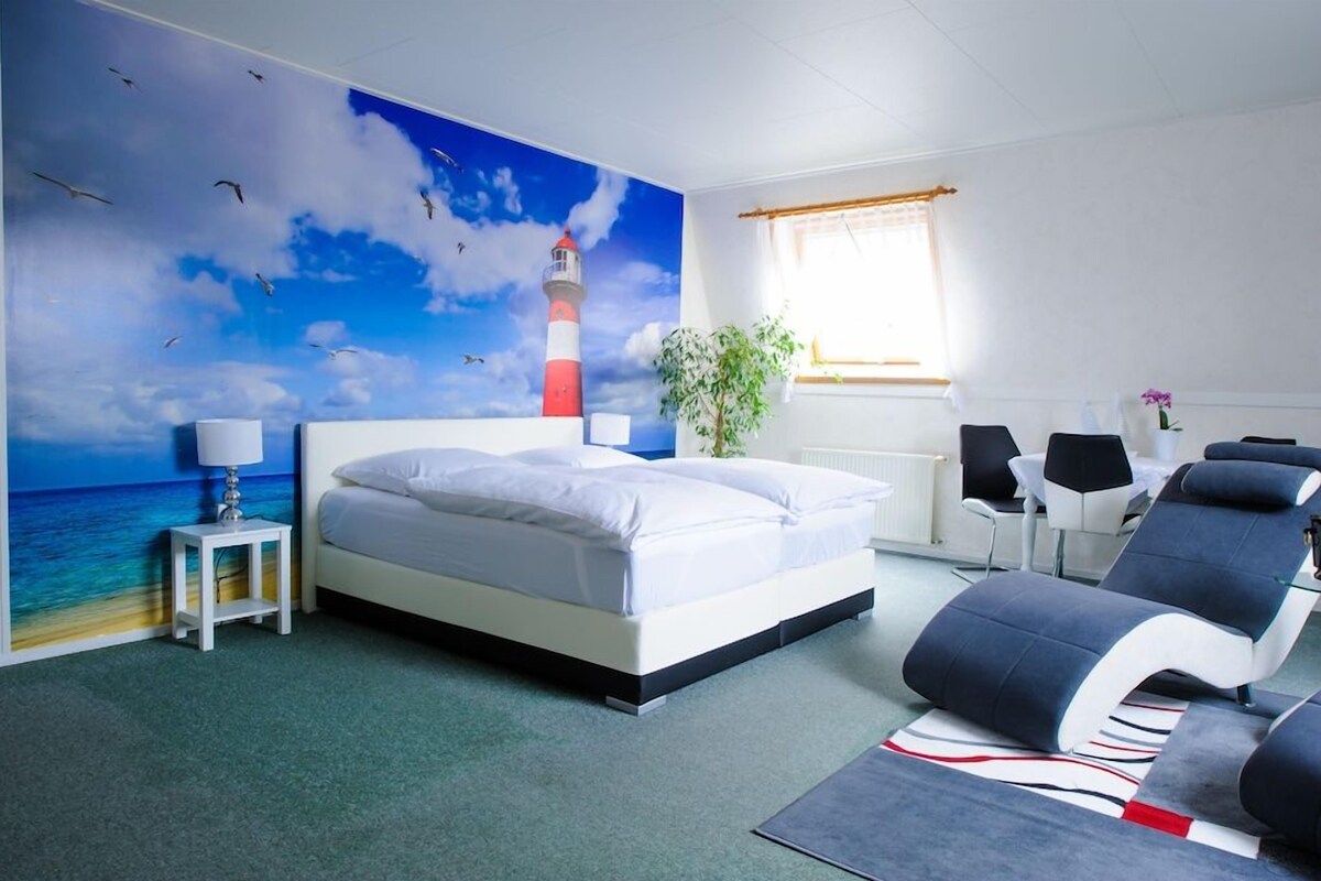 A hotel room with a bed, a lounge chair, a small table, a lamp, a plant, and a mural of a lighthouse.