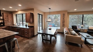A modern kitchen with dark wooden cabinets, a large island, and a dining area with a table and chairs. There is a sofa and a small table with a lamp in the living area. - Bend River Retreat (Bend)