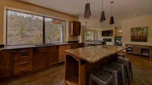 A modern kitchen with wooden cabinets, a marble island, and a large window overlooking a forest. - Bend River Retreat (Bend)