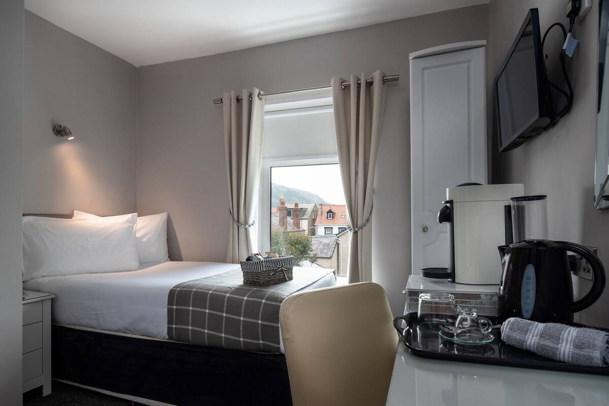Habitación de hotel con cama, escritorio, cafetera, tetera y ventana con cortinas.
