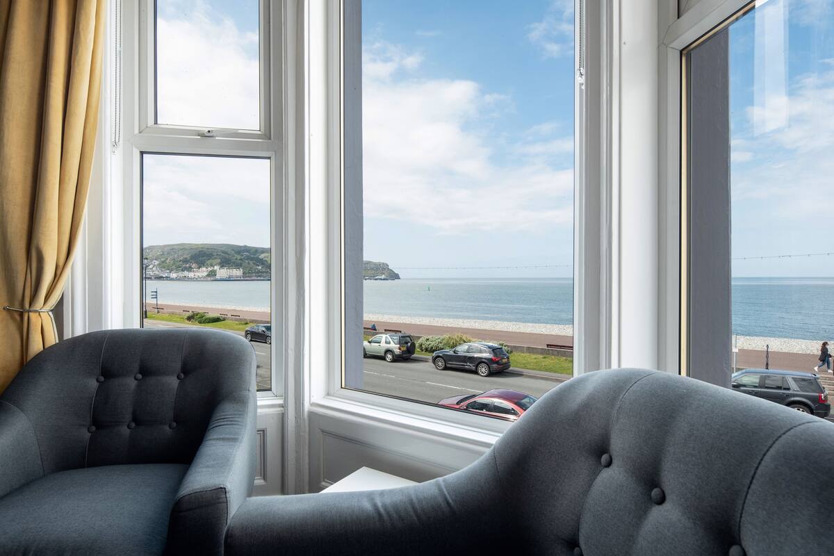 Una habitación con dos sillones grises frente a un ventanal con vistas a una carretera costera, coches y una playa.