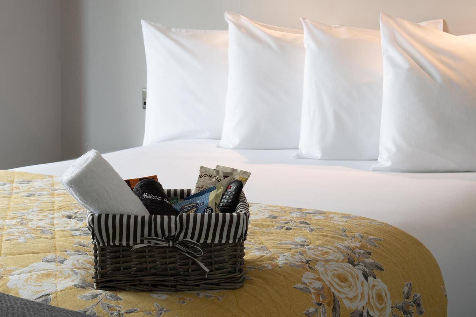 A neatly made bed with white pillows and a yellow floral bedspread. In front of the bed, there is a wicker basket containing a folded towel, a remote control, and some currency notes.