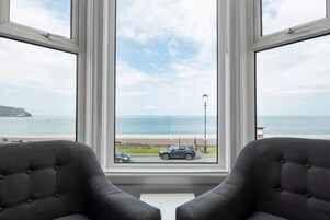 Habitación con un amplio ventanal que da a una playa y al mar, con dos sillones grises tapizados y una mesa blanca.