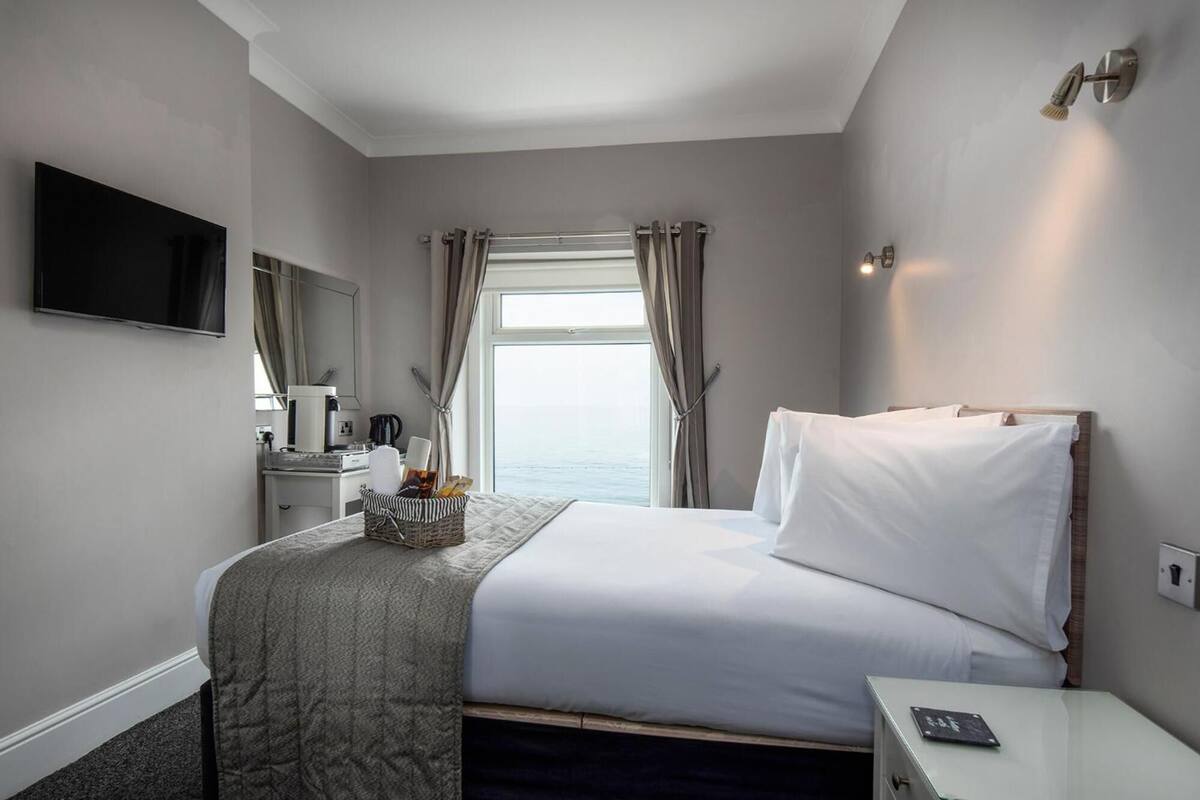 Habitación de hotel con cama, mesita de noche, televisor, ventana con cortinas y vista al mar.