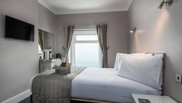 A hotel room with a bed, a bedside table, a TV, a window with curtains, and a view of the sea.