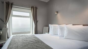 A hotel room with a large bed, white bedding, a view of the sea through a window, and a desk with a chair.