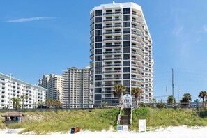 Un hôtel de grande hauteur doté de nombreux balcons, situé près d’une plage où se trouve un poste de surveillance des sauveteurs et un panneau indiquant la proximité d’un spot de surf.