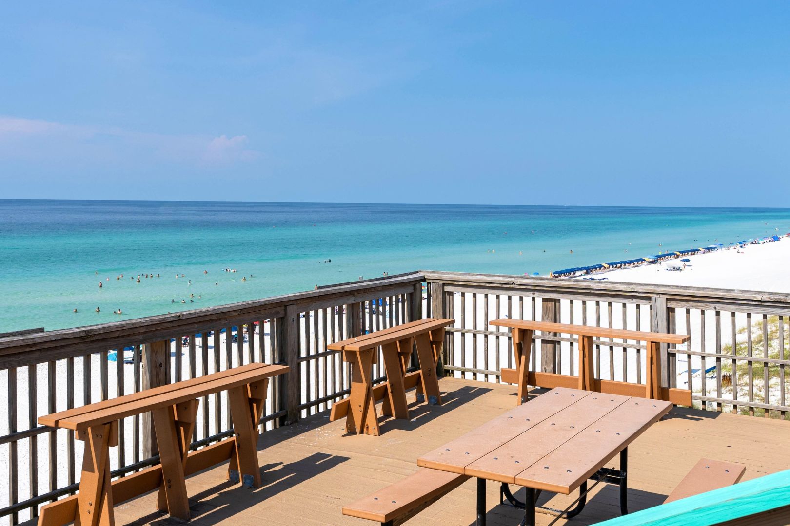 Une terrasse en bois avec des tables de pique-nique, donnant sur une plage où des gens se baignent et prennent des bains de soleil.