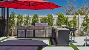 Een barbecue-opstelling met een grill, oven en zithoek, omringd door een witte schutting en bomen.