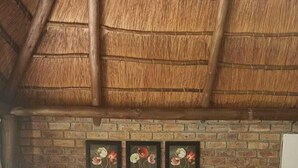 A bed with a grey blanket and a rolled towel, a brick wall with three flower paintings, and a wooden ceiling. - Pepe Piatto (Dipaleseng)