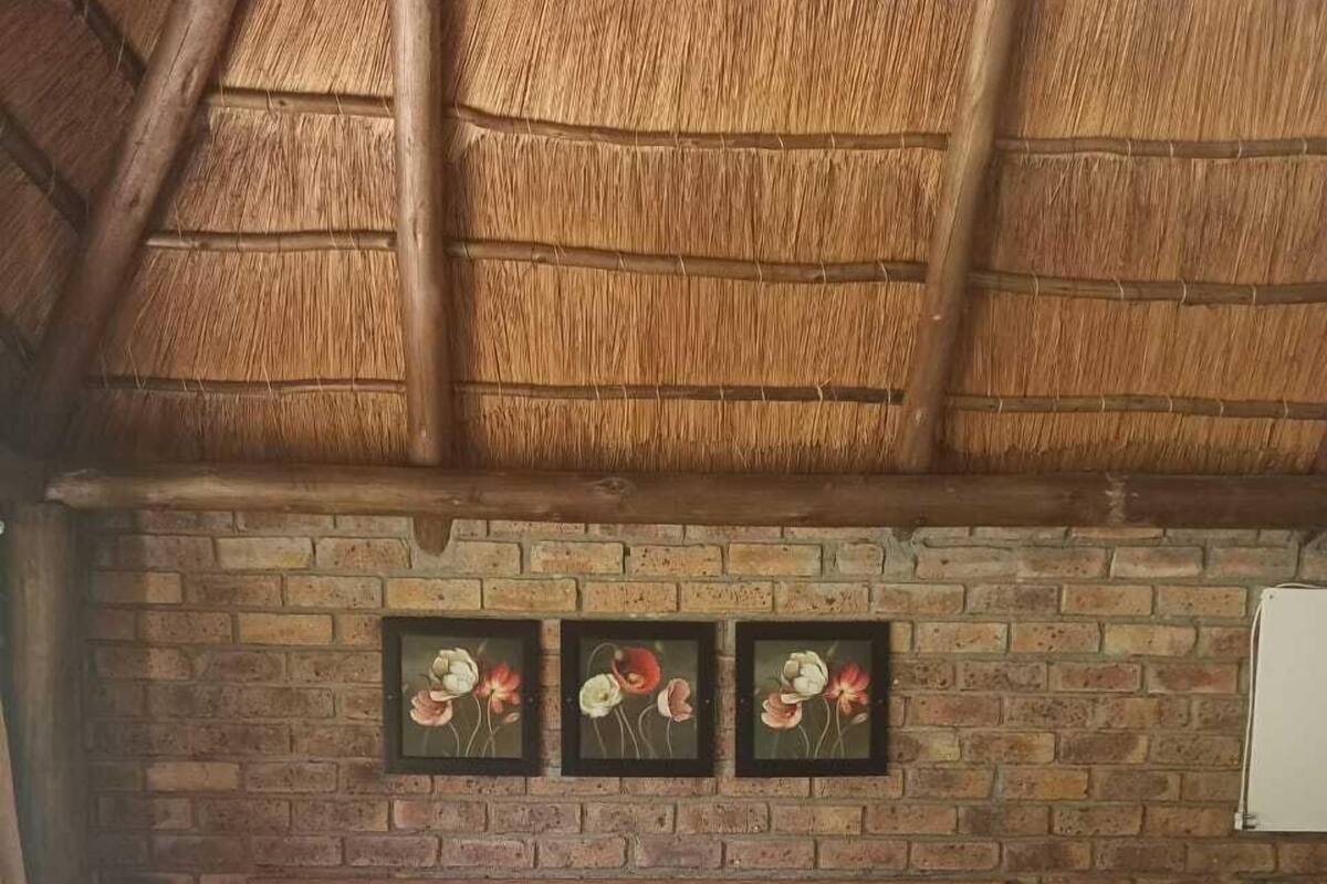 A bed with a grey blanket and a rolled towel, a brick wall with three flower paintings, and a wooden ceiling.