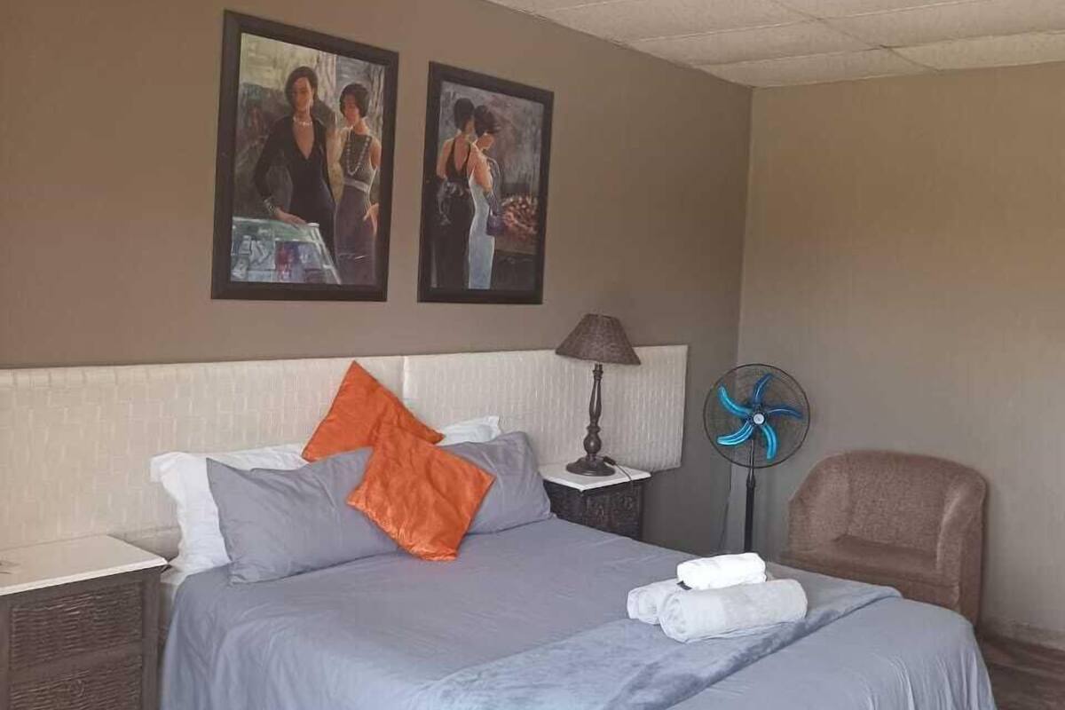 A hotel room with a bed, two wall-mounted paintings, a bedside table with a lamp, a fan, and a chair.