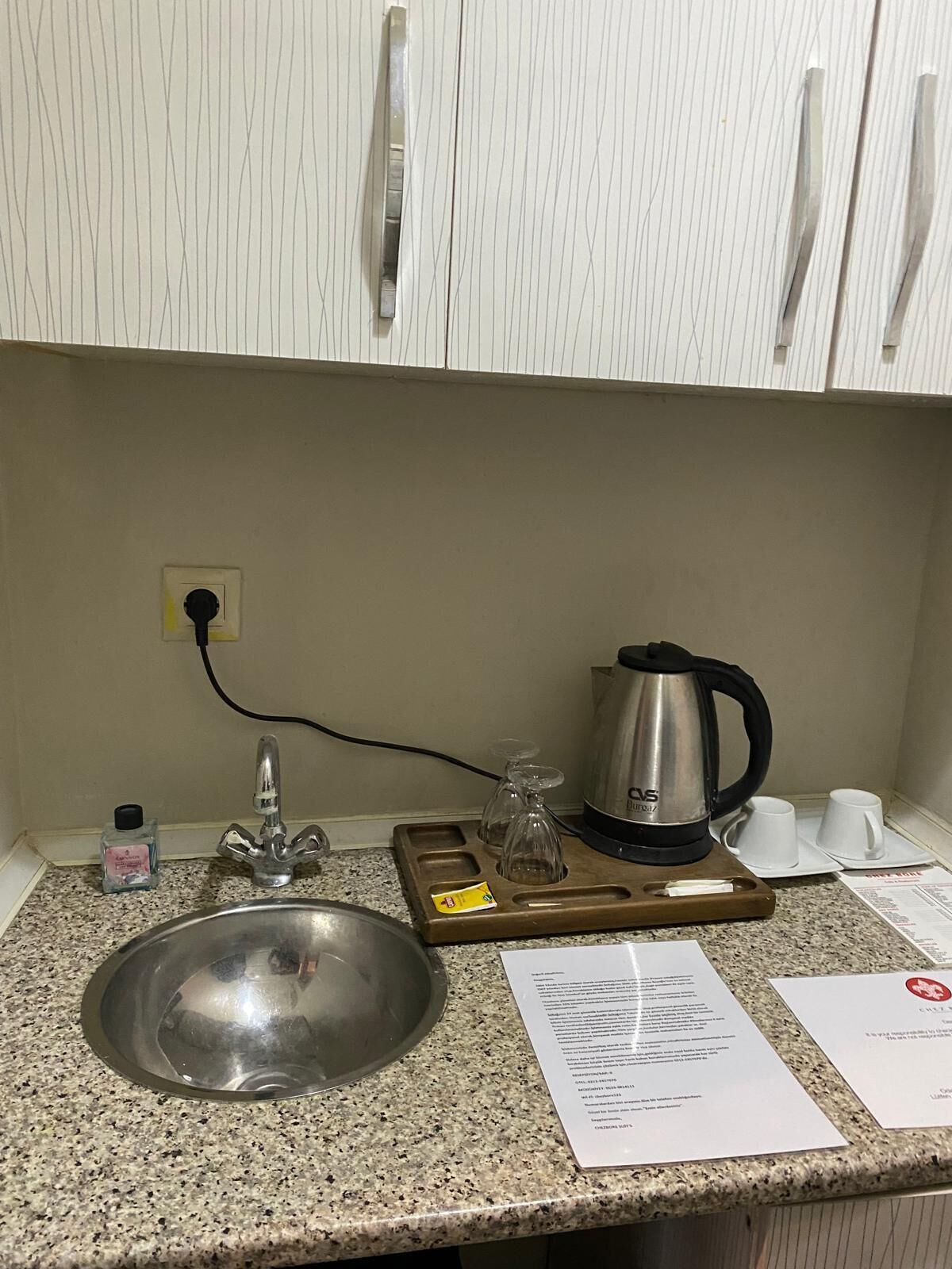 Cozinha com pia de aço inoxidável, chaleira e bandeja com copos. Há um papel no balcão e uma garrafa de sabonete líquido.