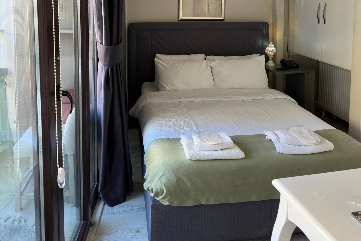 A neatly made bed with white linens, a bedside table with a lamp, and a view of the outdoors through a sliding glass door.
