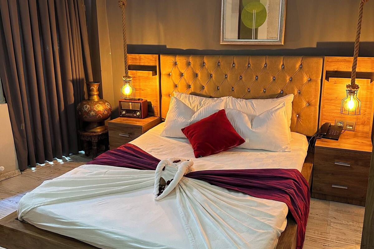 A neatly made bed with a white comforter and a red pillow, a bedside table with a lamp, and a framed picture on the wall.