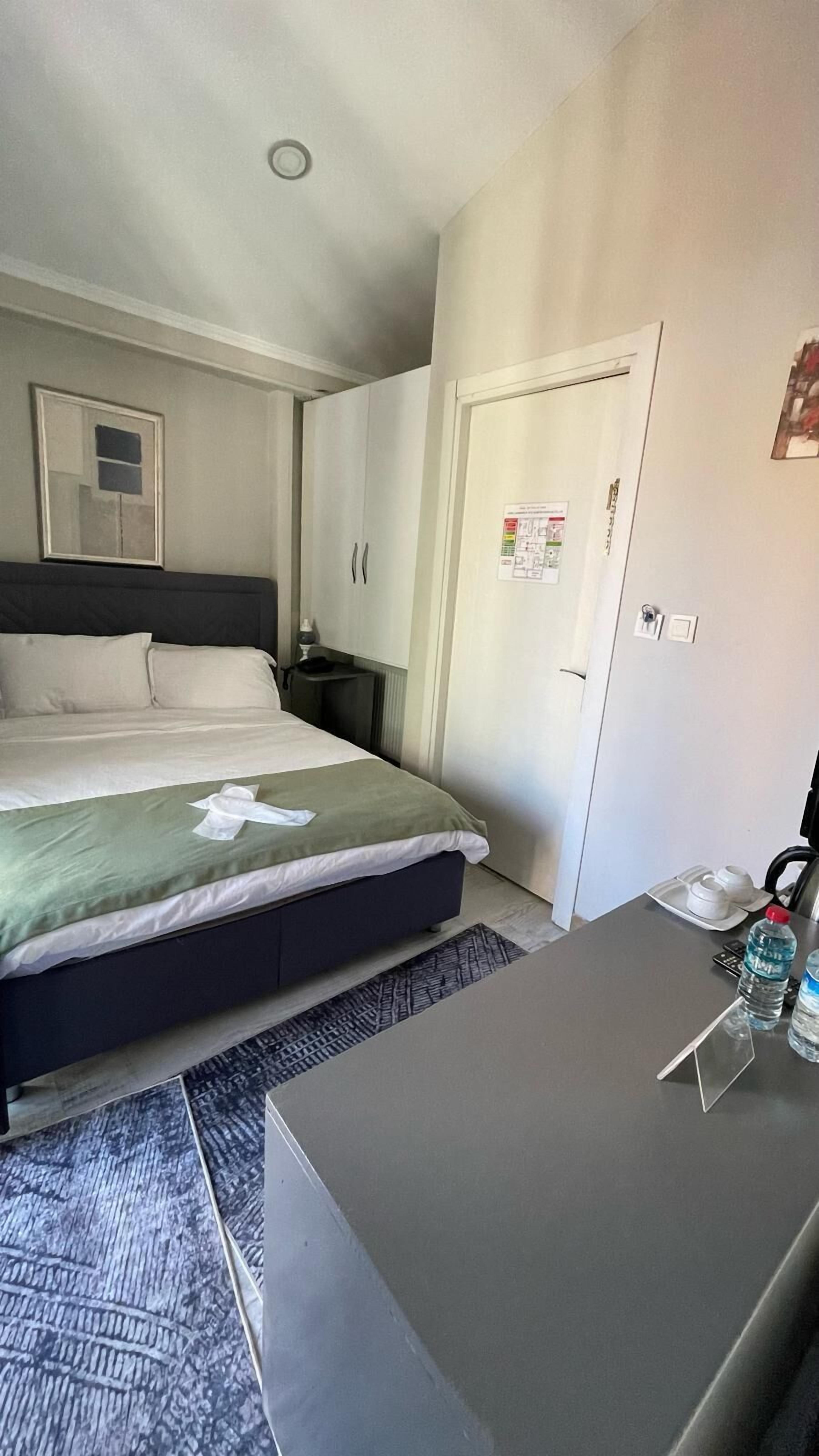 Une chambre d’hôtel avec un lit, un bureau, une chaise, une table de chevet, un miroir, une porte ornée d’une affiche et une petite table sur laquelle se trouvent une tasse et une bouteille.
