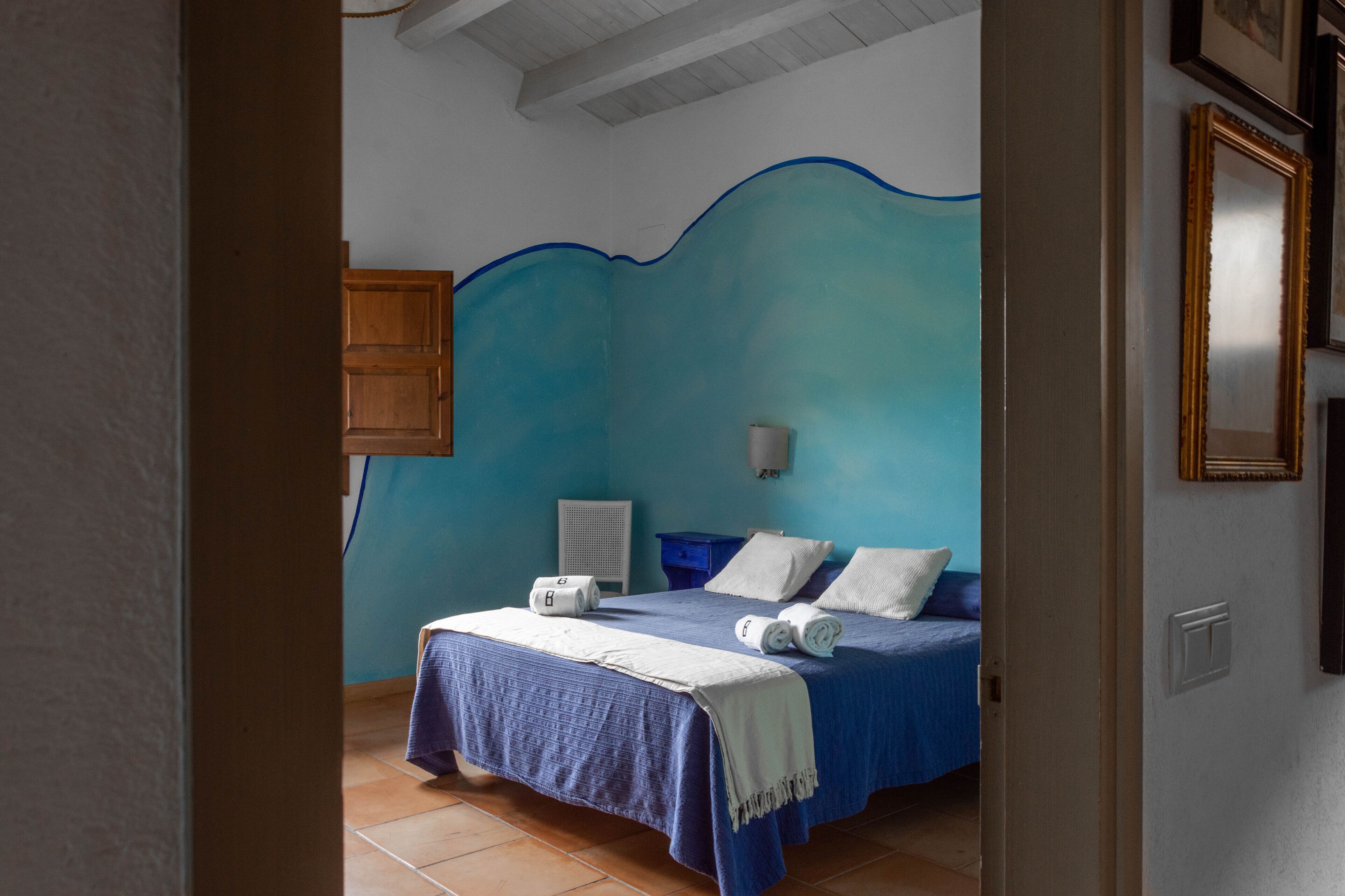 Un dormitorio con cabecera azul, una cama con ropa blanca, una mesita de noche y una lámpara de pared.