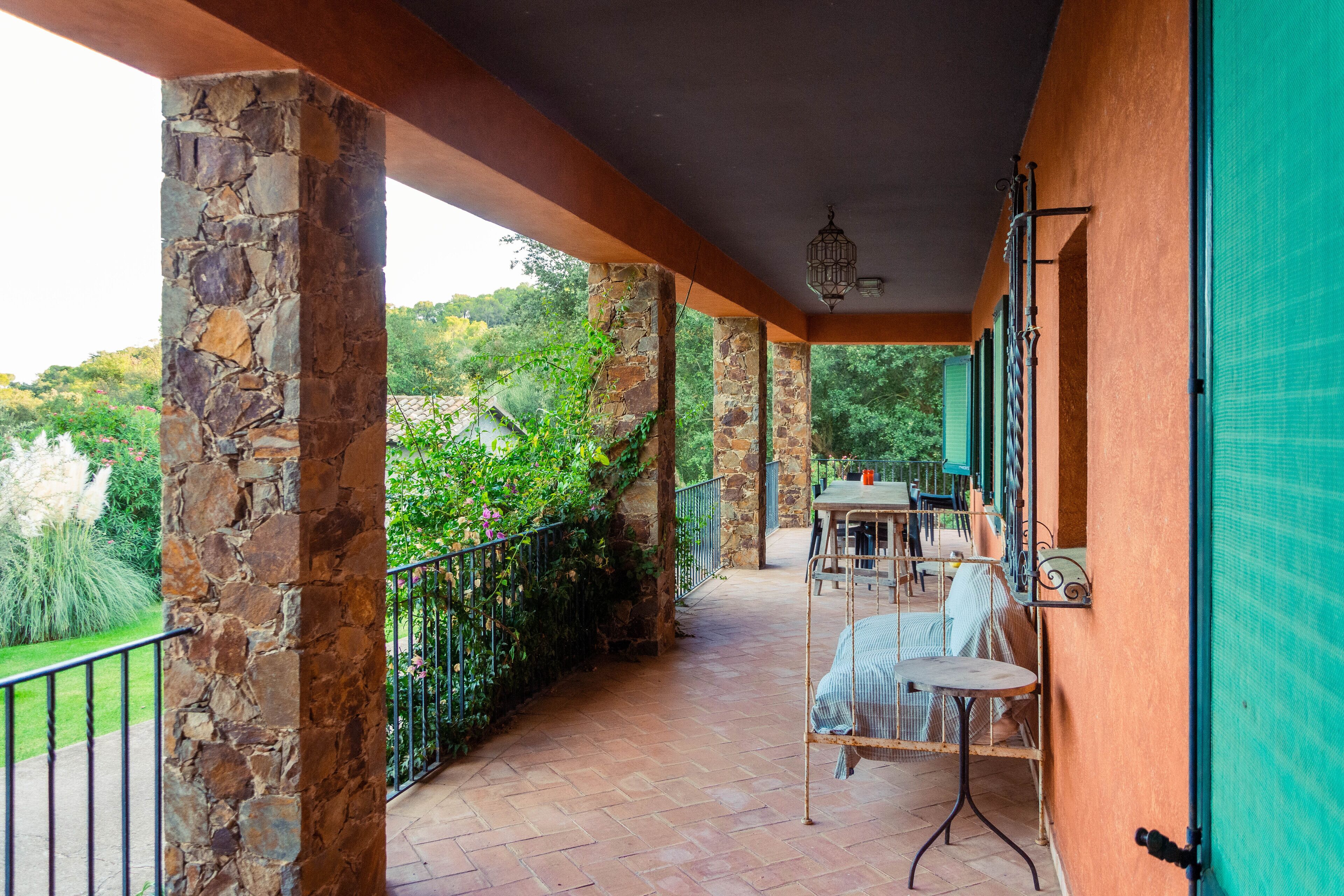 A covered outdoor patio with stone pillars, a table with chairs, and a view of greenery and trees.