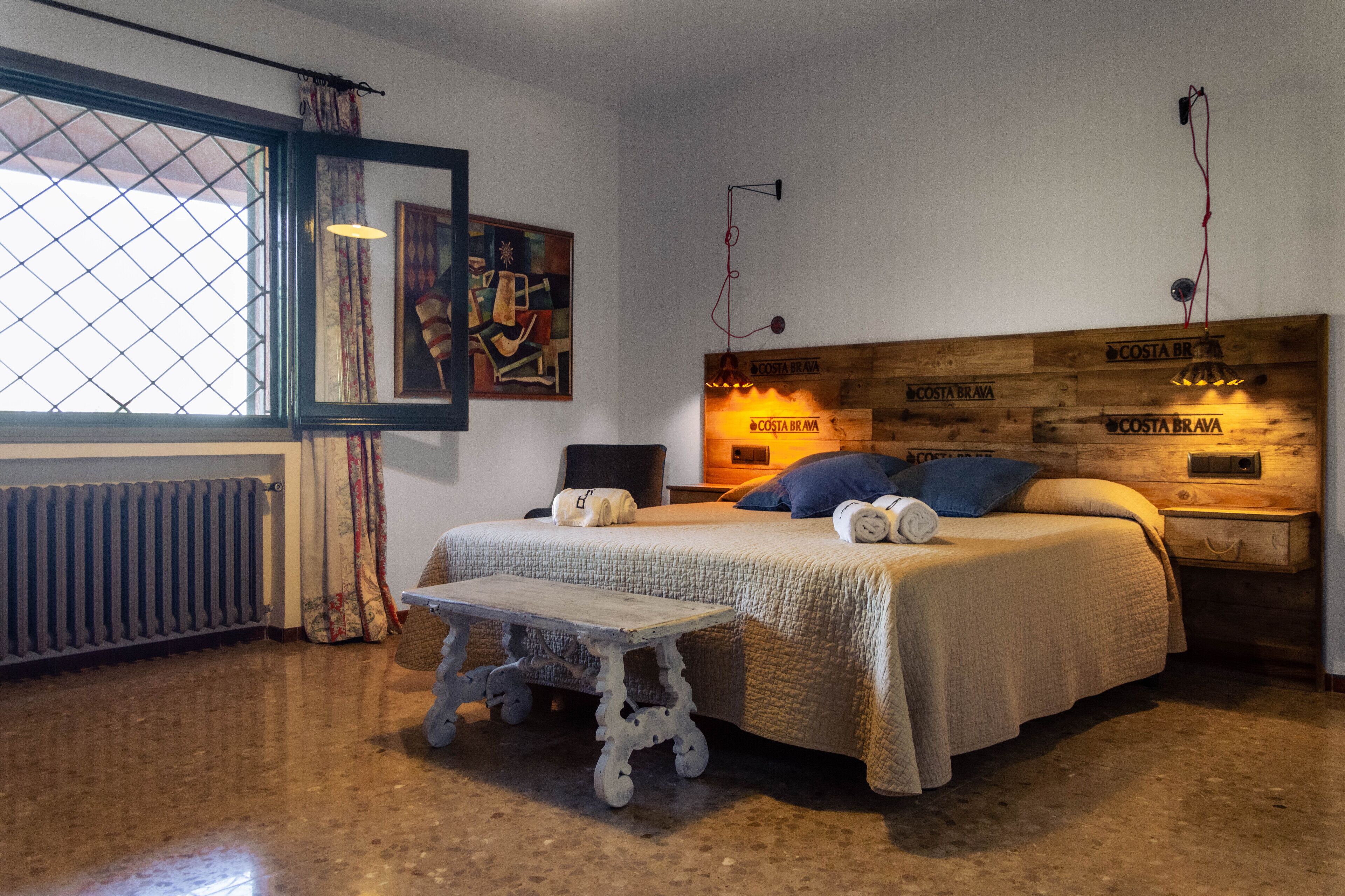 A bedroom with a wooden headboard, a bed with pillows, a small table, a radiator, and a window with curtains.
